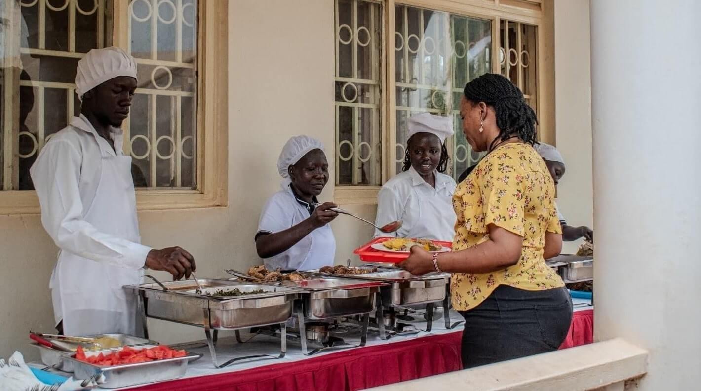 Members of Wot Monye (in white aprons) serve meals at the Re:BUiLD livelihood resource center. Wot Monye started as a savings group, it later started catering services for events. The group is one of the service providers for IRC events.